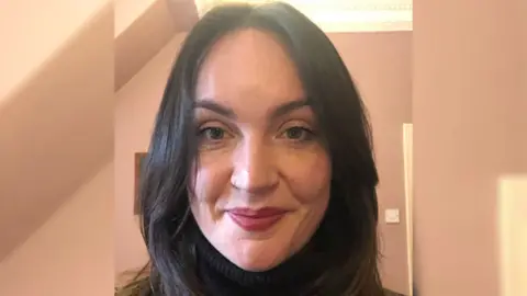 Sarah Hossak Teacher Sarah Hossak smiles to the camera. She has dark shoulder length hair and dark eyes. She is sitting in a pink-coloured room.
