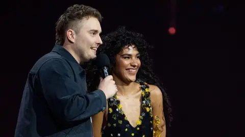 Getty Images Sam Fender and Olivia Dean in smart clothes on stage together