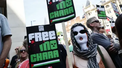 A person wearing a white balaclava and a headscarf holds a poster that says 'Free Mo Chara' . Other people in the background also hold placards