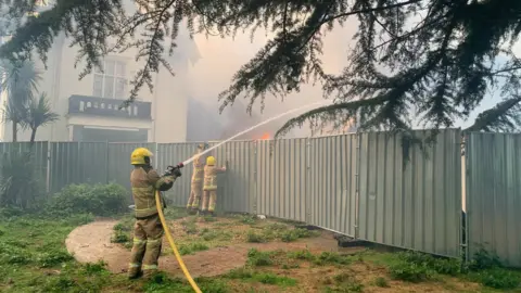Shropshire Fire and Rescue Service A firefighter aims water from a hose over a high metal fence. On the other side of the fence can be seen buildings, flames and smoke.