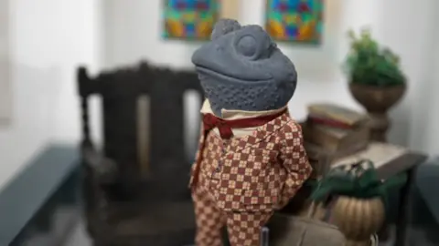 Prop of Toad in a red-and-white check suit and red bow tie standing in a quaint living room set. A wooden chair is in the background with books and plant pots stacked on a side table.