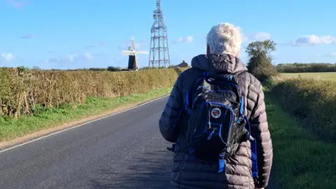 Daniel Charlton March marcher walking towards Over Windmill. The marcher has short white hair, is wearing a grey jacket and has a blue and black backpack on their back. They are on the right side of a tarmacked road, with a verge and hedge on either side of the road. 