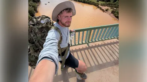 A man with a hat, grey jumper and black shorts, and with a rucksack on his back, takes a selfie while on a bridge over a brown-coloured river. He has placed his right foot on top of a football.