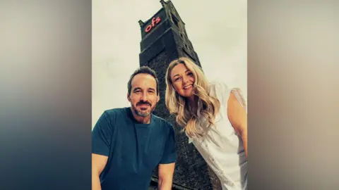 OFS Will and Maxine Morgan, who run the Old Fire Station in Carlisle. They are crouching down towards the camera, with the old watchtower from the former fire station behind them, with the OFS sign illuminate on top.