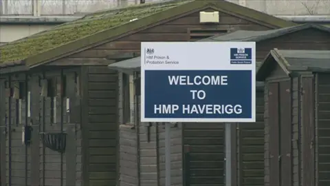 A blue and white sign saying "Welcome to HMP Haverigg". It is in front of hut-style buildings painted brown outside the main gate of the prison.