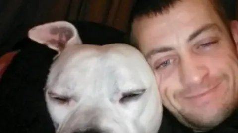 A man with short brown hair and stubble is next to a white Staffordshire bull terrier-type dog.