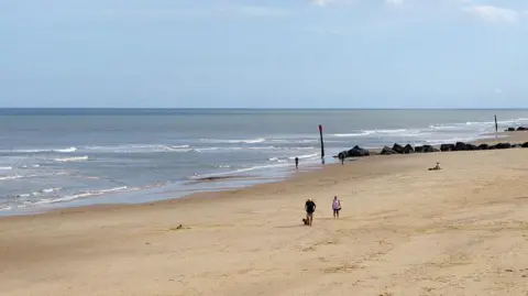 Stephen McKay/Geograph A couple and a dog can be seen walking along Waxham beach on a sunny day, with a row of rocks acting as sea defences visible in the background
