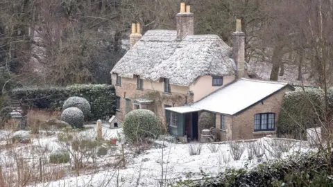 Thatched cottage standing in gardens with woodland behind - a sprinkling of snow can bee seen on the ground and the roof of the cottage.