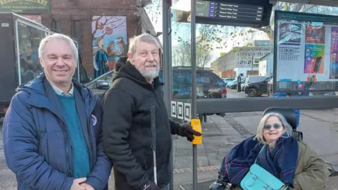 Two men stand by a bus stop alongside a woman using a wheelchair. All three are smiling towards the camera. The man in the middle is holding a walking stick and has his other hand on a large yellow button attached to the side of the bus shelter.