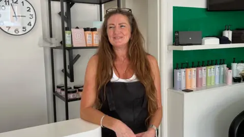 Kate Bradbrook/BBC A woman stands inside a hair salon. She is looking away from the camera as she smiles. She has long brown hair, glasses resting atop her head, and she wears a black apron with a white vest underneath. 