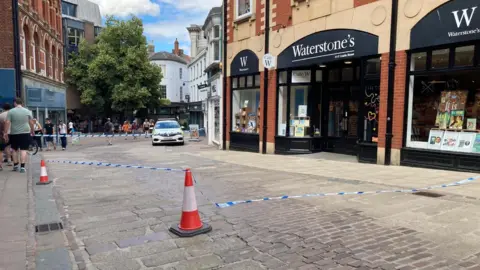Blue police tap attached to cones and a building has cordoned off a section of a city street in Norwich, outside a Waterstone's store, which has black and white branding, with books in the windows on display. A police car can be seen in the background next to the cordon, as people walk past it. 