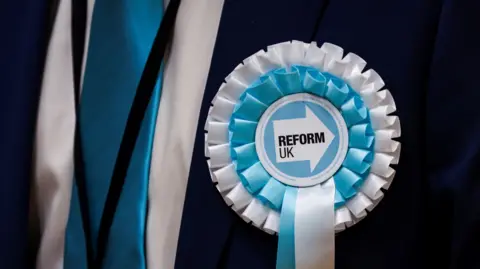 A close up of a blue and white Reform UK rosette on a dark suit jacket. A white shirt and blue tie can also be seen.
