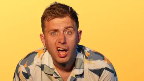 Mark Westgate Martin Westgate wearing a colourful shirt and sneering as part of a promotional photo. He has short brown hair, blue eyes and is standing in front of a yellow background