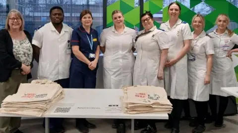 NHS Humber Health Partnership Six nurses, featuring five women and one man and wearing white nurse's uniforms, are standing behind a table at an apprenticeship event. Also pictured with them is a senior nurse in a blue nurse's dress and a woman in civilian clothing.