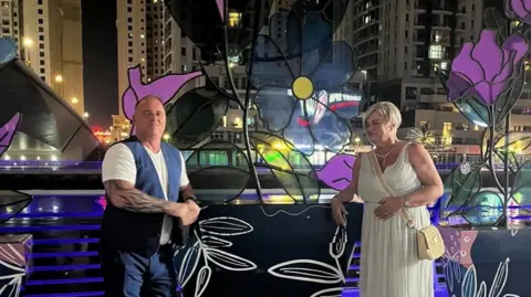 Ray wearing a white t-shirt and blue waistcoat is leaning against a railing in Dubai and smiling at the camera at night. To the right Mel has short blonde hair and is wearing a white, Grecian-style dress and is also leaning against the railing. She is looking at him.