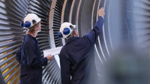 Two males stand inside a giant, circular metal construction which could resemble a very large fan with slats in the metal. They are wearing white hard hats and blue jumpsuits and one man is pointing at something. 