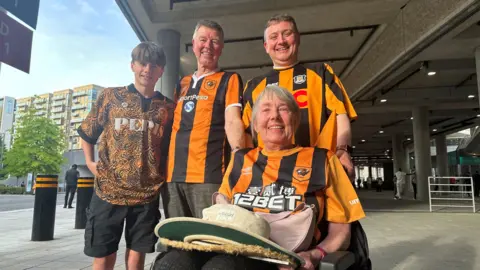 BBC/ Matt Dean A family all wearing Hull City shirts, a woman with grey hair is seated in a wheelchair, behind her are three men all with brown hair and smiling.