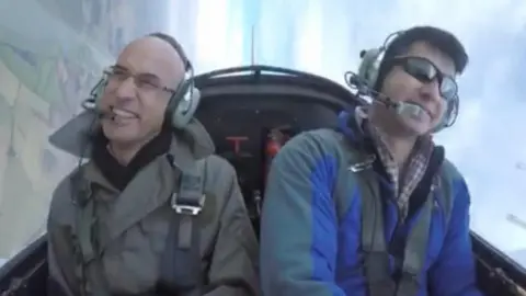 Amrik Sandhu A photo of two men in a plane they are both smiling and have got headphones on and plane seatbelts. They are both wearing jackets, one is blue and one is grey. One man has got sunglasses on and the other one has sunglasses on. 