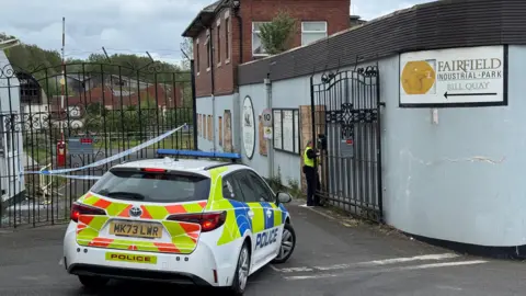 A police car is parked beside a sign for Fairfield Industrial Park. There is an officer behind a large, black metal gate. In the background there are buildings.