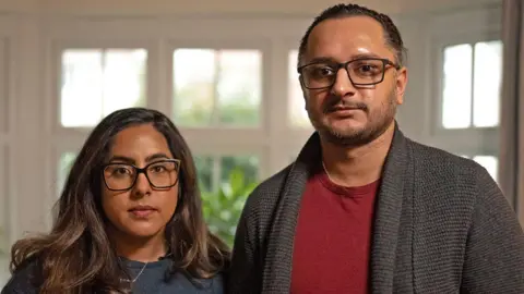 MARTIN MCQUADE/BBC A couple stand next to each other with serious expressions. The light shines through the window behind them. 