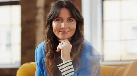 A woman with long brown hair and a blue suit, she is sitting on a yellow sofa and is resting wit her hand on her chin