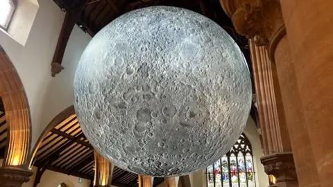 The Moon sculpture hanging from the ceiling of the cathedral. It is grey and features many craters. There are terracotta coloured columns in the foreground and stained glass windows in the background.