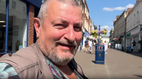 Dean smiles at the camera. He is the main focus and in the foreground. He has a short grey beard and wears a grey gilet and shirt. Blurred behind him is a street of shops.