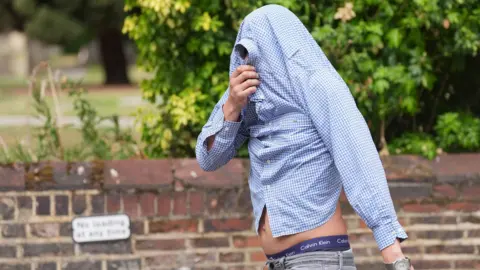PA Media A man wearing a checked blue buttoned down shirt but he has pulled it up over his head to cover his entire face and has a small gap of fabric to see out of. He is also wearing grey jeans. He is walking along the pavement and behind him is half a brick wall and trees and bushes.