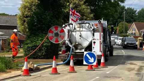 A white tanker truck is parked next to a green hedge. There is a row of cars on the opposite side of the road. There are orange cones behind the vehicle and a man wearing orange florescent overalls. There are pipes running from the read of the tanker.