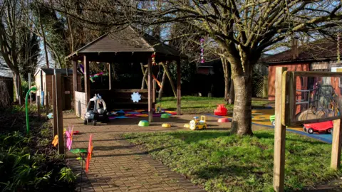 City of Wolverhampton Council A garden with a paved path, grassy areas and trees. An outdoor seating area with pitched roof is at the end of the path and small children's toys lie around the garden including a ride-on truck and car.