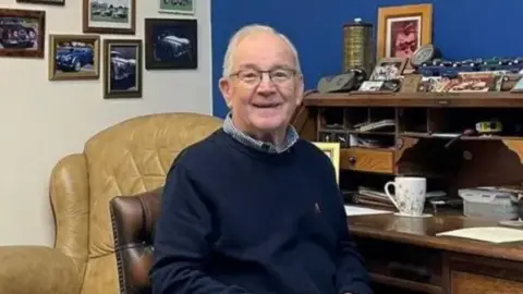 A man with grey hair and glasses is sat at a desk, smiling to the camera. Behind him are pictures of old cars