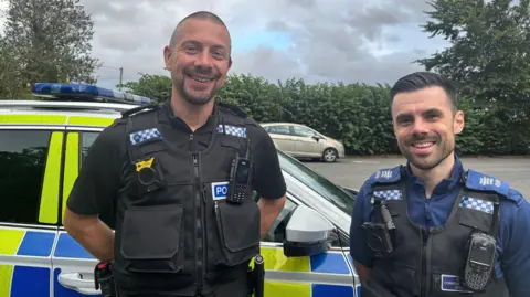 police officer is standing in front of a police car. he has short brown hair and facial hair, and is wearing a black police vest with a walkie talkie and camera. Next to him is a PCSO - he has long brown hair and short facial hair, he is wearing a navy blue shirt with a black vest over the top that has a camera and walkie talkie.