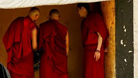Three monks stand in a row in red robes talking among themselves, their faces truned away from the camera