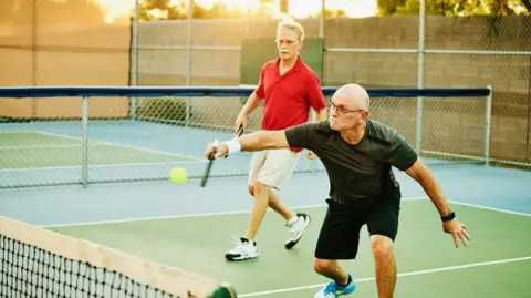 Getty Images Wide shot of a man hitting backhand shot at net while playing doubles pickleball on summer evening 
