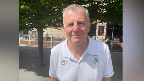 James Burridge/BBC Paul Stephens standing in St George's Square in Luton, wearing a white collared t-shirt with the Luton Town logo on the left and Umbro logo on the right. He has grey cropped hair and smiles at the camera. There are trees behind him and outdoor cafe seating.
