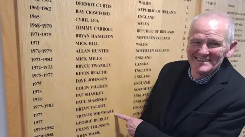 Craig Whymark An elderly looking man with little hair and wearing a black coat over a jump. He is pointing at a wooden wall board which has names of Ipswich Town players written on it.