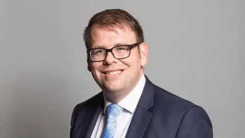 UK Parliament Young man with glasses in a navy suit jacket, white shirt, blue tie, smiles 