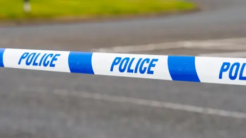 A section of white and blue police tape stretches across a paved road.