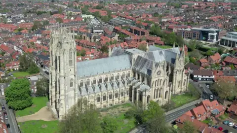 A wide-angle aerial shot of Beverley Minster, a large, gothic Cathedral-type building. Houses, green grass and trees surround. 