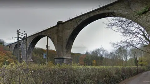 The stone Victoria Viaduct photographed from the path below. The viaduct has fencing with trees and shrubs surrounding its foundations.