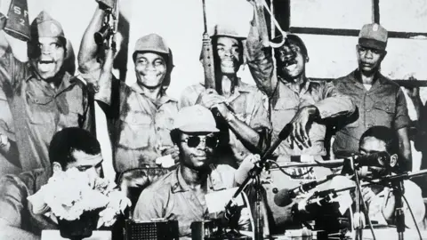 Eugene Shaw via Getty Images A black and white photo of Samuel Doe and his fellow coup makers, with some raising their guns, addressing a press conference.