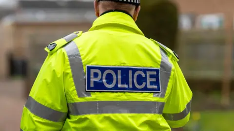 Stock image of the back of a police officer who is wearing a yellow high visibility police jacket. They appear to be walking towards some houses. 