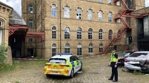Charles Heslett/BBC A police car with yellow and red stripes on it boot parked at a taped off scene in one section of the mill.