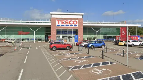 Google The tesco store is a long, rectangle shaped building with a box-like entrance with a sign above glass windows that says: "Tesco". In front of the supermarket is a car park with some cars parked in bays.