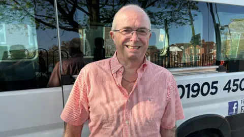Gavin Kermack / BBC A man with thinning white hair and glasses and a red and white pinstripe short-sleeves shirt stands in front of a white minibus, smiling at the camera.