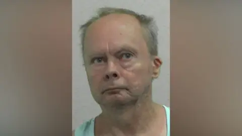 Mugshot of Henderson. He is clean shaven with a small chin and short balding grey hair.