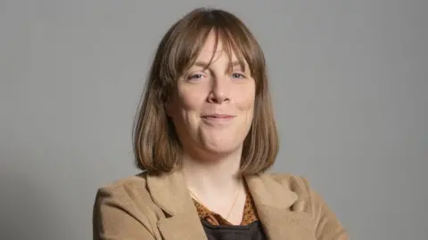 PA A woman in her 40s with a light brown bob smiles as she stands against a grey background in an official Parliament portrait. She is wearing a camel coloured coat and black top with an orange collar.
