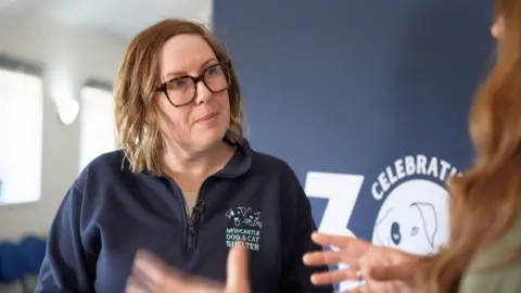 Anita talks to a BBC reporter. She has shoulder length brown hair and glasses and is wearing a blue fleece top with the charity's name on the breast.