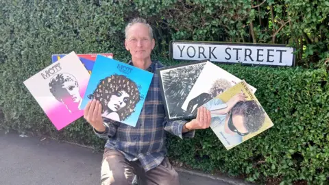 Duncan Kerr A man in a blue cheque shirt and brown trousers crouching down holding some Ian Hunter and Mott the Hoople albums