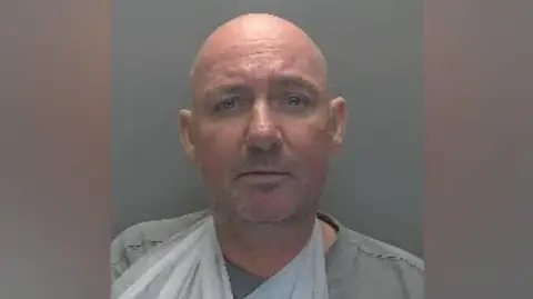 Cambridgeshire Police A police custody image of Peter Corrigan. He is wearing a grey T-shirt and a medical sling on one arm. He is standing in front of a grey wall.